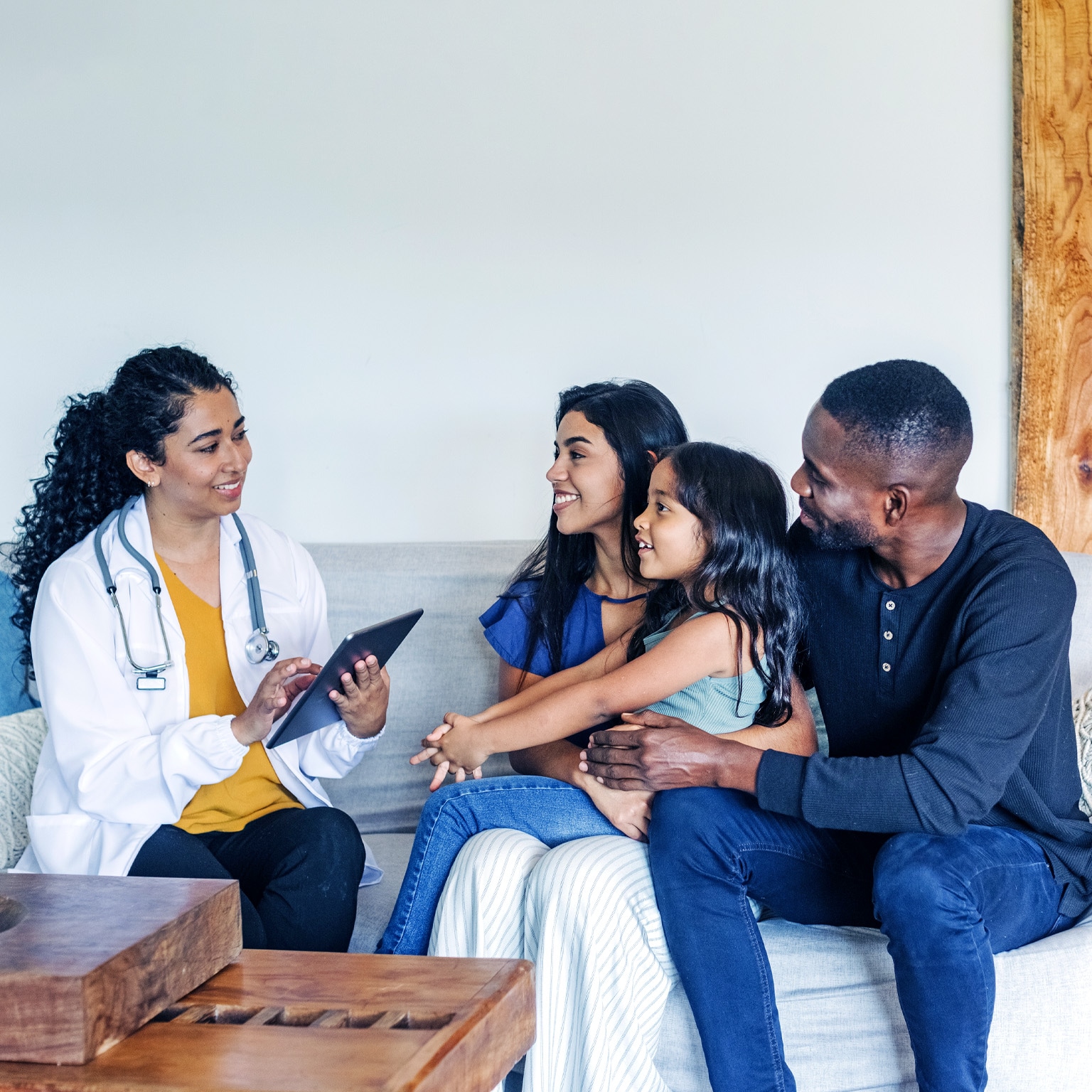 A hispanic doctor sitting on a couch talking to a mixed-race family during a home visit.