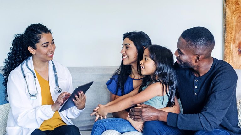 A hispanic doctor sitting on a couch talking to a mixed-race family during a home visit.