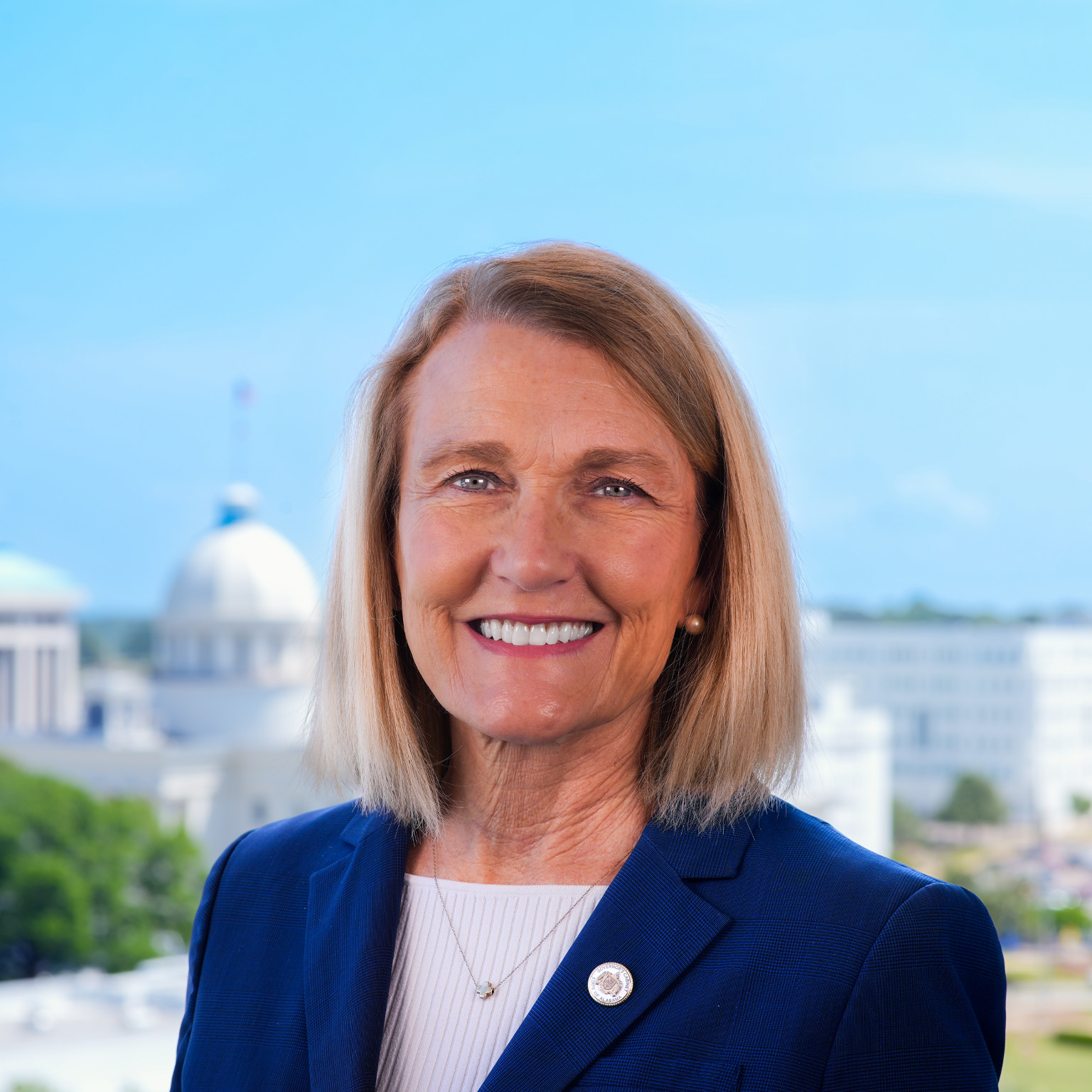 Ellen McNair with shoulder-length blonde hair smiles at the camera, wearing a blue blazer and light-colored top. Behind her, a softly blurred cityscape featuring a domed building and other urban structures under a clear sky.