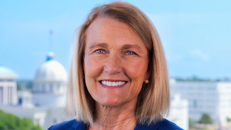 Ellen McNair with shoulder-length blonde hair smiles at the camera, wearing a blue blazer and light-colored top. Behind her, a softly blurred cityscape featuring a domed building and other urban structures under a clear sky.