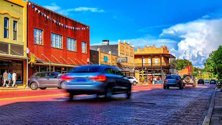 Cars and pedestrians are moving by on a busy main street in rural America with window-lined with shops and a cobblestone roadway