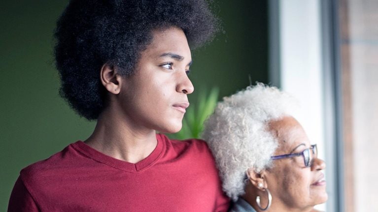 A young African American man stands beside his grandmother, his arm resting gently on her shoulder as they both gaze thoughtfully out of a window.