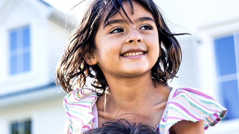 Young girl riding on her fathers shoulders in front of suburban home