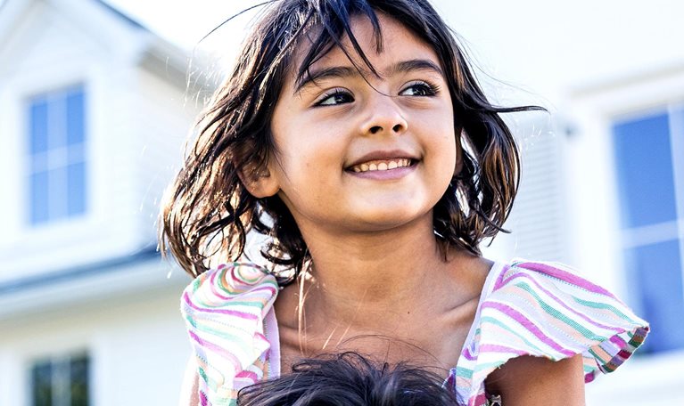 Young girl riding on her fathers shoulders in front of suburban home