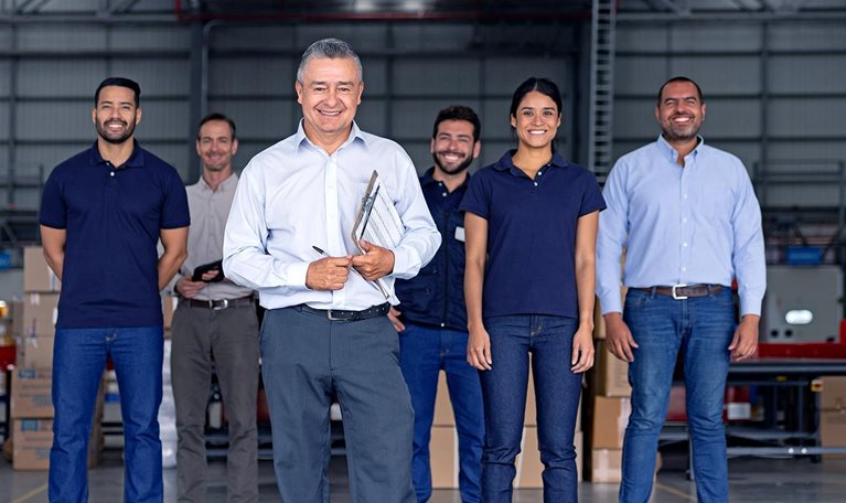 A man inside a warehouse leading a group of Latin American warehouse employees; he is holding a clipboard and the team is scattered behind him but all are smiling and looking at the camera