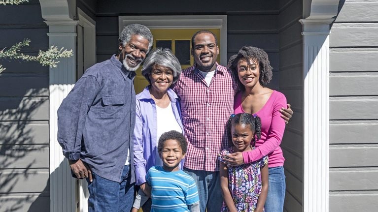 Three generations of a Black family pose together and smile outside their home.