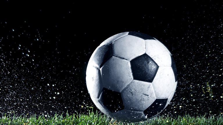 A black-and-white soccer ball sits on wet grass as water and turf spray outward, suggesting recent impact or motion.