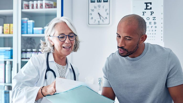 A doctor and a patient sitting together in a medical exam room, reviewing documents and discussing information in a folder. The setting includes medical cabinets, charts, and equipment, creating a calm, professional healthcare environment.