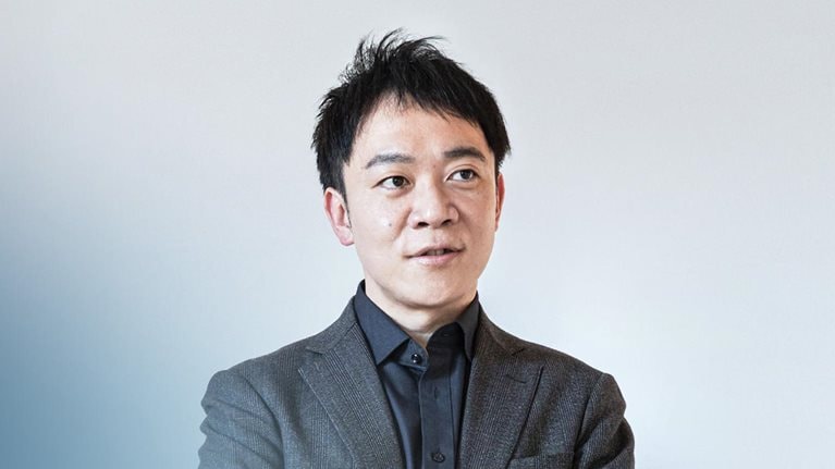 Portrait of Shinpei Kato in a gray suit against a light-gray background and with soft lighting. Kato turns his gaze away from the camera and looks off thoughtfully into the distance.