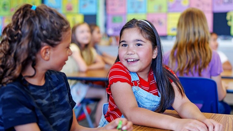 An elementary school girl sits with a friend at a desk in their classroom and talks cheerfully during class.