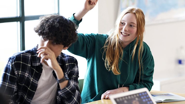 A group of students sitting at a classroom table, with a red-haired student in a green hoodie smiling and raising her hand while others, including one with a laptop, also participate.