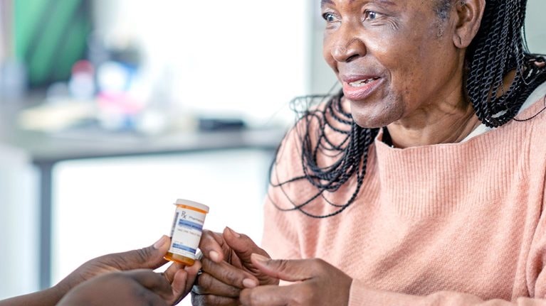 A senior woman sits across the desk from a nurse practitioner as they discuss her medications. She is dressed casually and looking at the nurse as she listens attentively. The nurse is dressed in blue scrubs and has a headscarf on as she holds out the medication and informs the woman about how to take the new medication.
