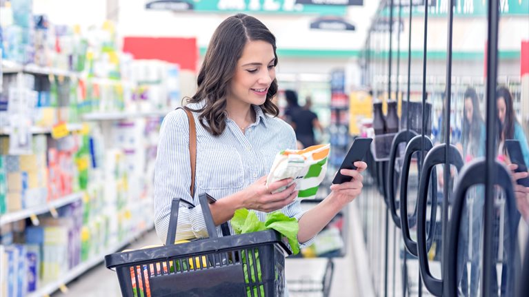 Confident young Hispanic woman holds smart phone as she reads a nutrition label on a bag of frozen vegetables. She is holding a shopping basket filled with healthy foods.
